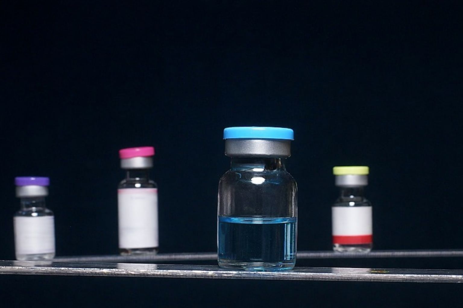 Små glasflaskor med olikfärgat innehåll och lock på ett mörkt underlag.